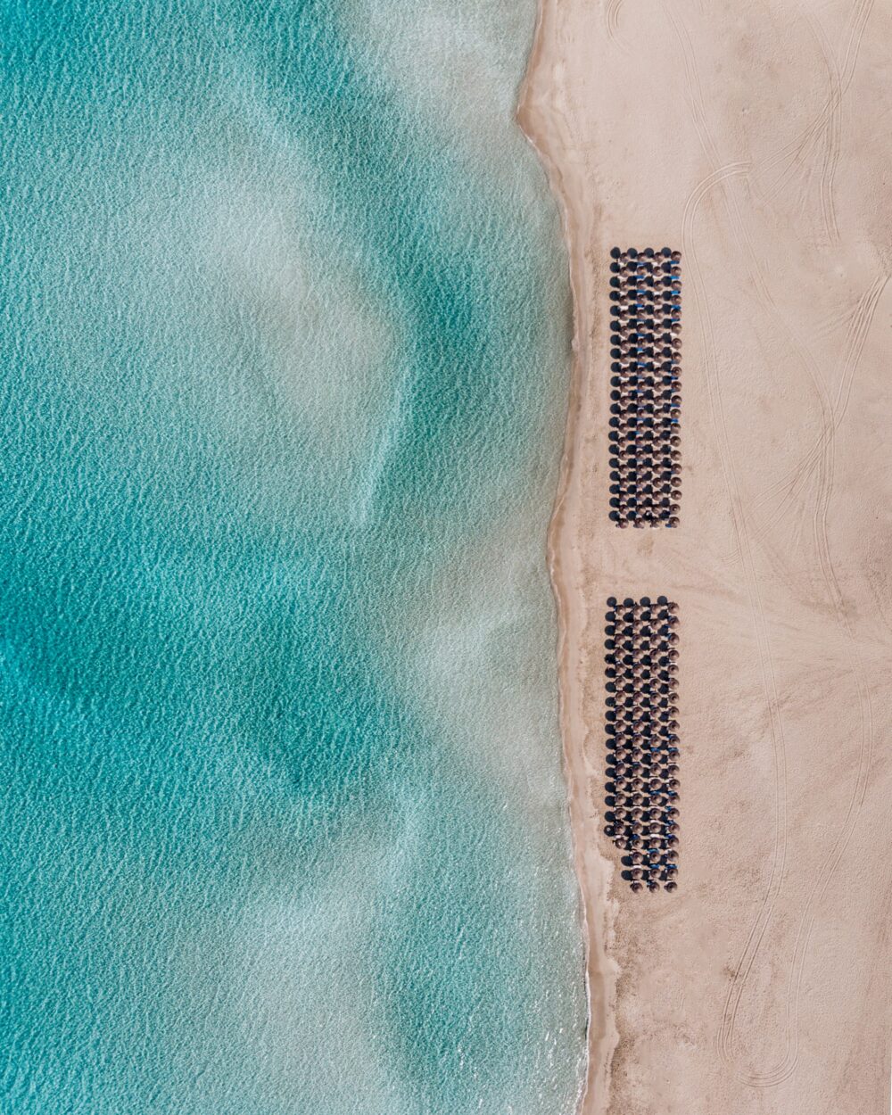 Aerial view of turquoise sea meeting a pale sandy beach lined with orderly rows of dark round umbrellas.