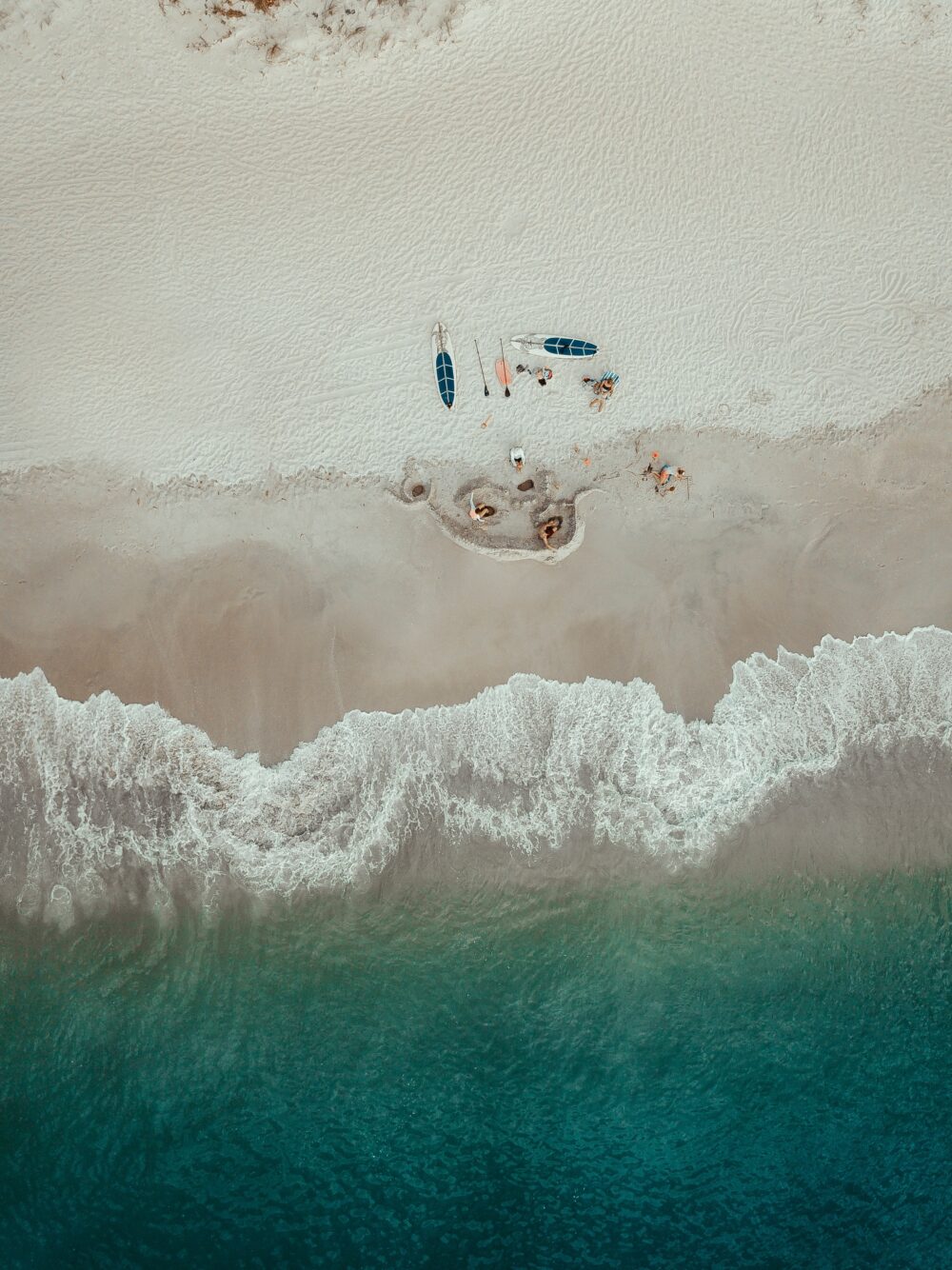 Aerial view of a pale sandy beach with paddleboards, people and surf at the shoreline.