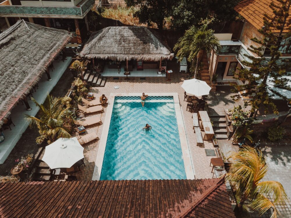 Aerial view of a rectangular hotel pool with turquoise water, loungers, white umbrellas and a thatched-roof pavilion.