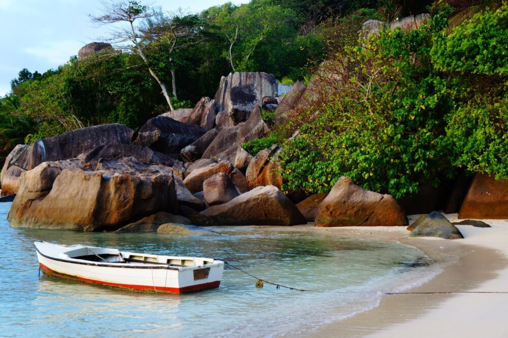 Small white boat anchored in shallow turquoise water beside large rocks and dense green coastal vegetation.
