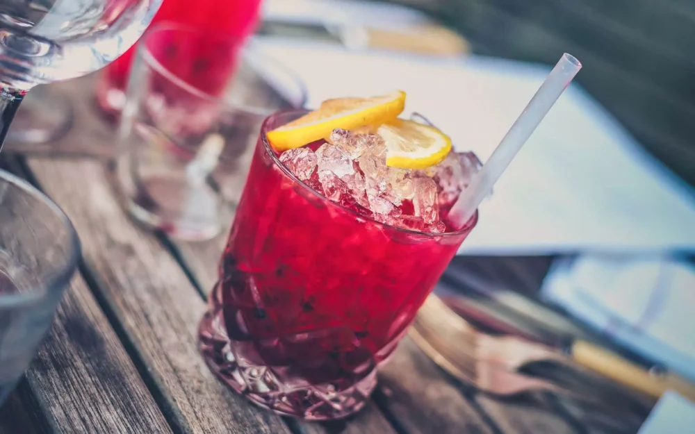 Red crushed-ice cocktail in a cut-glass tumbler with lemon slices and a straw on a wooden table.